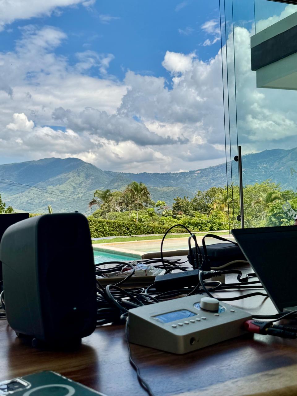 Interface de audio, monitores de estudio y laptop montados frente a un ventanal con vista a las montañas.